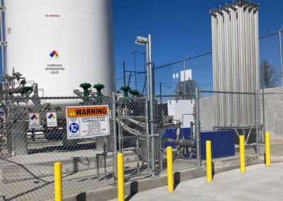A fenced area contains industrial hydrogen equipment, including tanks with warning signs about flammable liquid hydrogen. Yellow safety bollards line the fence, and the area is free of pedestrians.