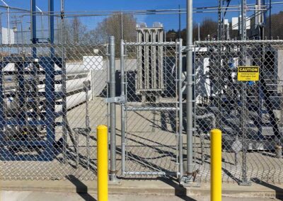 A fenced-off utility area with metal pipes and equipment behind a chain-link fence, two yellow bollards in front, and a caution sign warning of high voltage on the gate.
