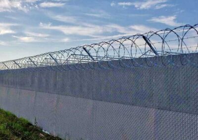 A tall chain-link fence topped with spirals of barbed wire stretches alongside a grassy area under a partly cloudy blue sky.