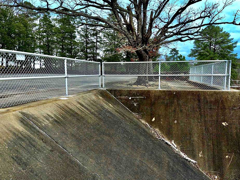 A chain-link fence runs along the edge of a concrete embankment near a road, with a large leafless tree and evergreen trees in the background under a partly cloudy sky.