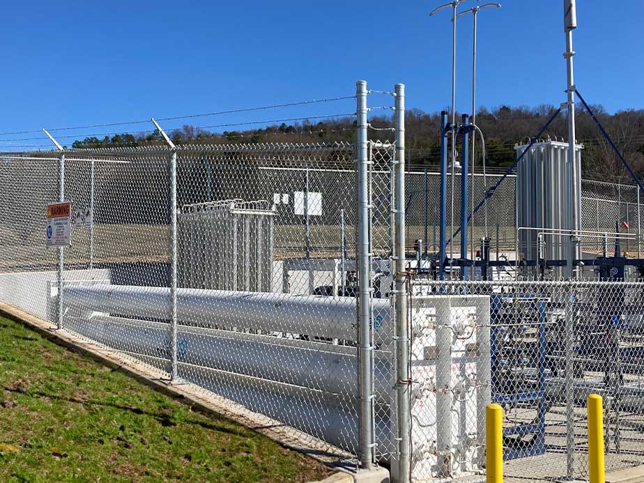 A fenced industrial facility with pipes, tanks, and tall structures, set on a concrete pad. Warning signs are posted on the fence. Grassy ground and a forested hill are visible in the background under a clear sky.