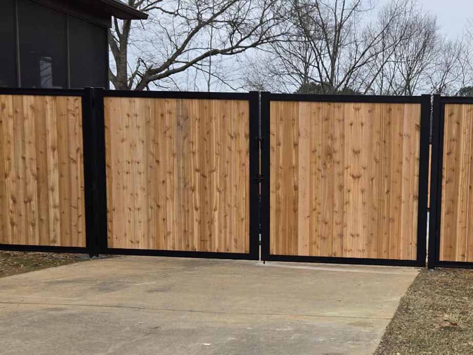 A wooden privacy fence with vertical planks and black metal frames encloses a yard, with a gate in the center and bare trees visible in the background.