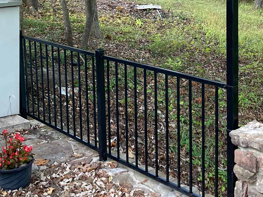 A black metal gate with vertical bars separates a stone patio with fallen leaves and a potted plant from a grassy, wooded area.