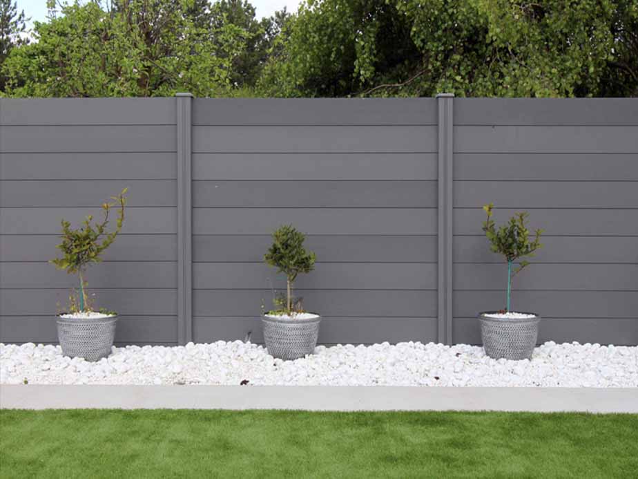 Three small potted trees are placed on white stones in front of a tall, gray fence, with green grass in the foreground and leafy trees in the background.