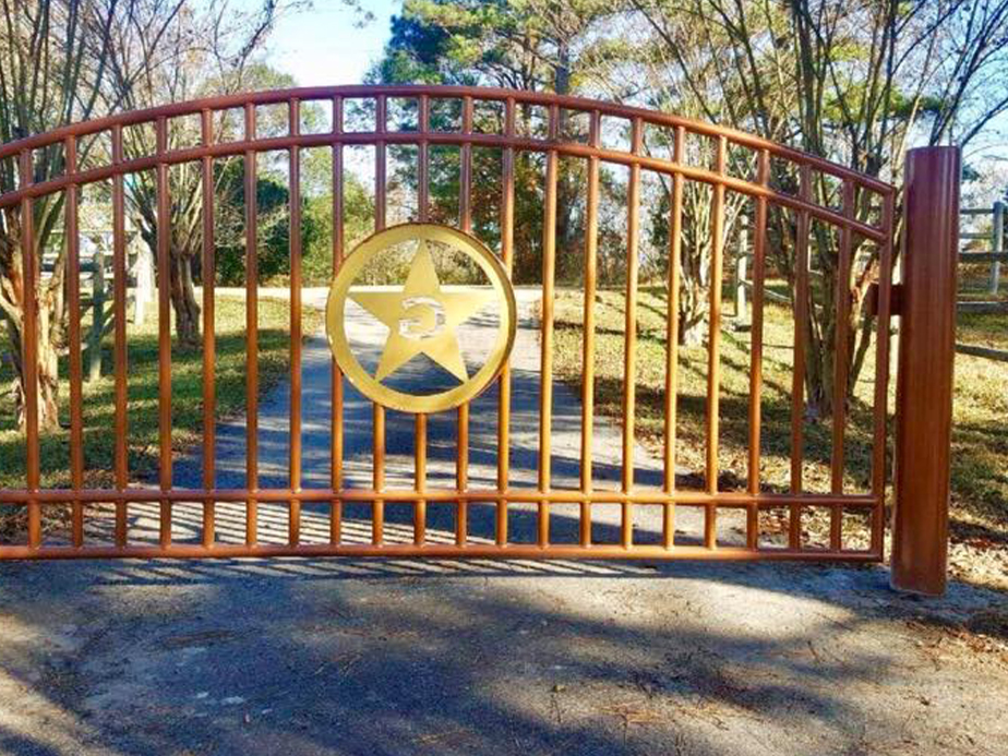 A brown metal gate with a large gold star emblem in the center blocks a paved driveway lined with trees and grass on a sunny day.
