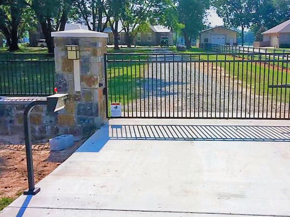A black metal sliding gate with a stone pillar and mailbox stands at the entrance of a gravel driveway leading to a house surrounded by grass and trees on a sunny day.