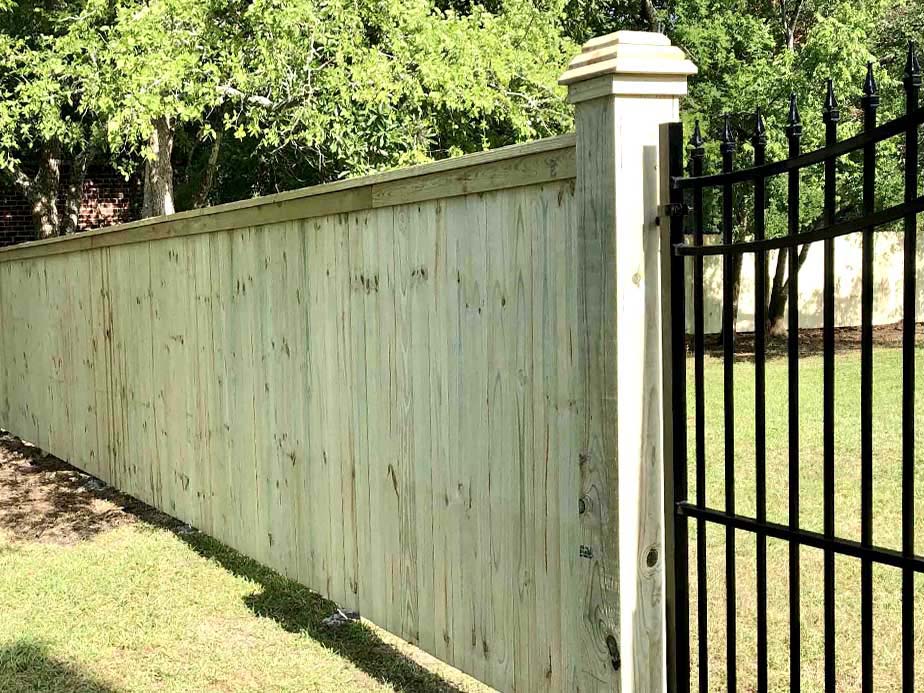 A tall wooden privacy fence with a decorative post stands next to a black metal gate, separating a grassy yard with trees and a brick wall in the background.