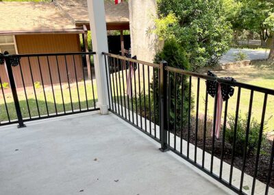 A porch with black metal railings decorated with black and white polka dot bows and American flags, overlooking a green yard with trees and a wooden shed in the background.