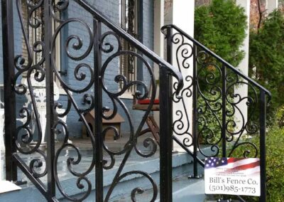 A decorative black wrought iron railing lines the steps of a blue-painted porch. A sign nearby reads Bills Fence Co. (501) 985-1773 with an American flag design in the background.