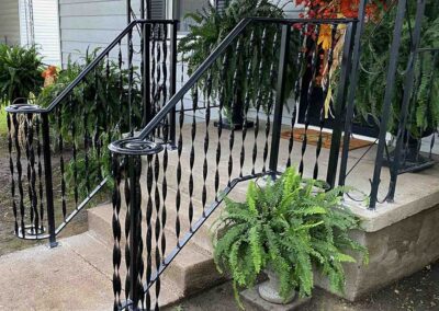 Black wrought iron railings with decorative twists line concrete steps leading to a porch. Lush green ferns and other potted plants decorate the steps and porch area.