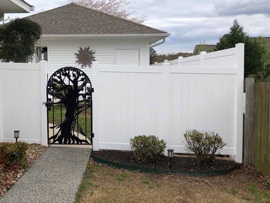 A white vinyl privacy fence with a black metal gate featuring a decorative tree design, leading to a house with a white exterior and neatly trimmed shrubs in front.