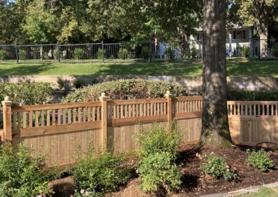 A wooden fence stands in a landscaped yard with bushes, flowering plants, and a large tree. In the background, there is a black metal fence, trimmed grass, and a white house partially visible through more trees.