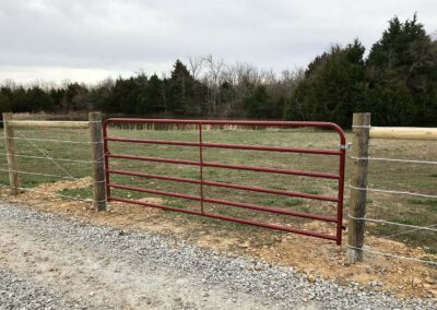 A red metal farm gate attached to wooden fence posts with wire fencing, set along a gravel road, with a grassy field and trees in the background under a cloudy sky.