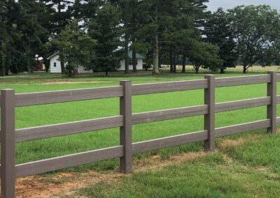 A wooden fence runs along the edge of a bright green grassy field with trees and a white house with a green roof in the background.