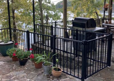 A patio with potted plants, a black metal fence surrounding a grill, and outdoor seating overlooks a wooded lake. The scene features stone flooring and lush green trees in the background.
