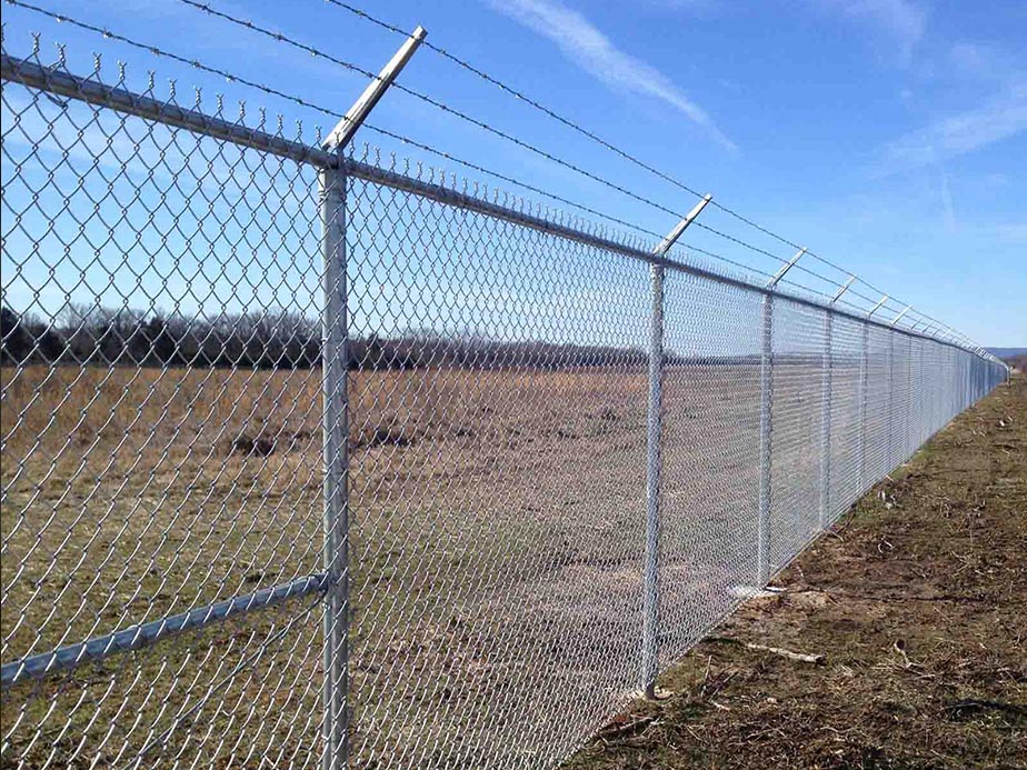 A long chain-link fence topped with barbed wire runs along a grassy, open field under a clear blue sky. The fence extends into the distance, dividing the landscape.