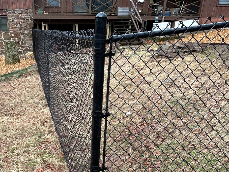 A black chain-link fence runs through a yard with patchy grass, leading toward a wooden house with a stone chimney and visible stairs in the background.