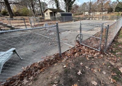 A fenced outdoor area with a closed gate, fallen leaves on the ground, and a covered swimming pool. Trees and a small building are visible in the background.