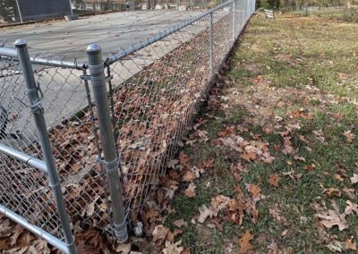 A metal chain-link fence runs alongside a leaf-covered grassy area, separating it from a concrete surface, possibly a covered pool. Fallen autumn leaves are scattered on the ground.