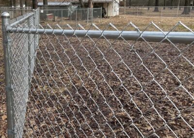 A close-up view of a metal chain-link fence enclosing a yard covered with dry leaves, with more fencing, utility boxes, trees, and a building visible in the background.