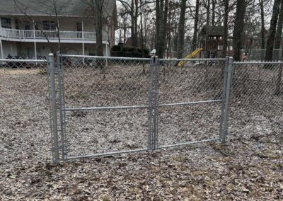 A chain-link fence with a double gate stands open, but there is no fencing on either side, making the gate nonfunctional. Behind the gate, there is a yard with scattered leaves, trees, and a play structure.