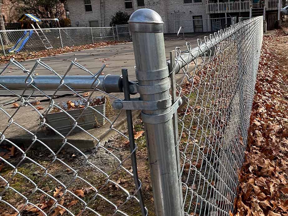 A close-up view of a metal chain-link fence gate with a latch, surrounded by fallen leaves. In the background, there is a playground slide and an apartment building.