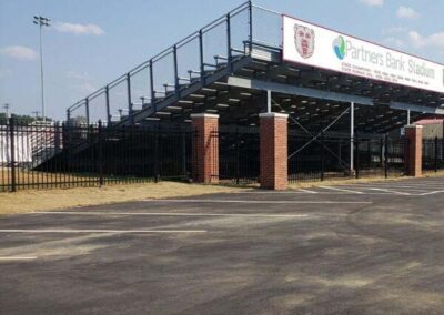 Bleachers at Partners Bank Stadium with a tall black fence and brick pillars in front, viewed from a parking lot on a sunny day. A sign for the stadium is visible on top of the bleachers.
