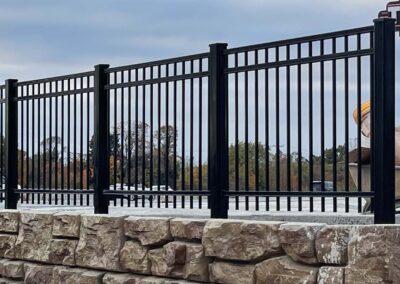 A black metal fence with vertical bars is installed on top of a beige stone wall. Trees and a cloudy sky are visible in the background.