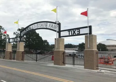Entrance gate to the Arkansas State Fair, with brick pillars, metal arch, flags, and signs reading TIX and EXIT. A fairground and domed building are visible in the background.