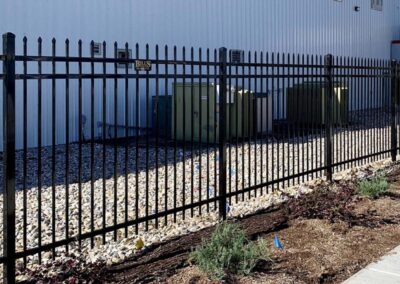A black metal fence with pointed tips encloses electrical utility boxes on a bed of rocks next to a white industrial building. There are some small plants and mulch in the foreground outside the fence.