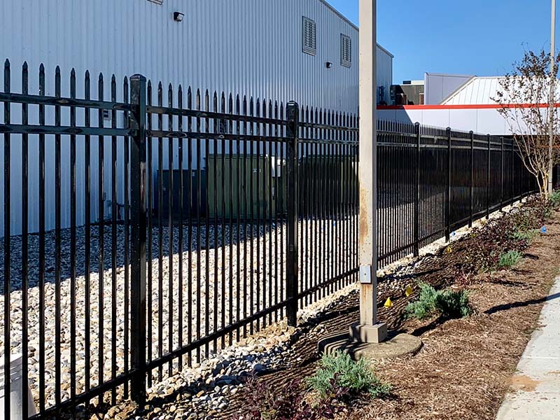 A black metal fence with pointed tops runs alongside a sidewalk and landscaped area, next to a white industrial building on a sunny day. Rocks cover the ground inside the fenced area.