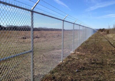 A long chain-link fence topped with barbed wire runs alongside a grassy, open field under a blue sky. The ground nearby is uneven with patches of dirt and sparse grass.