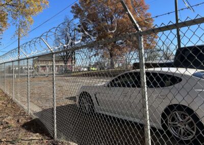 A white car is parked next to a chain-link fence topped with barbed wire. Behind the fence, trees with autumn leaves and utility poles are visible under a clear blue sky.