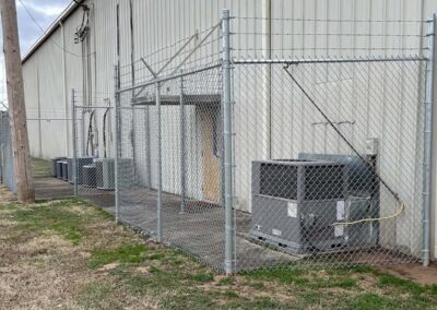 A chain-link fence encloses several outdoor HVAC units next to a large metal industrial building. The fenced area is on a concrete walkway, with grass and utility poles nearby.