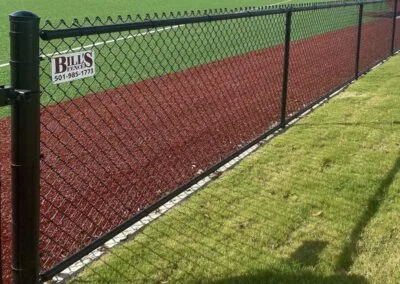 A black chain-link fence separates a green artificial turf field from a strip of red track and a grassy area. A sign on the fence reads “Bill’s Fence” with a phone number.