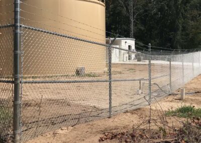 Chain-link fence with a bent section near the center, enclosing a sandy area with a large tan industrial tank and a small white building in the background. Trees are visible beyond the structures.