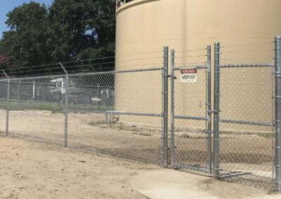 A chain-link fence with a locked gate and a KEEP OUT warning sign surrounds a large beige industrial tank. Trees and other fencing are visible in the background.