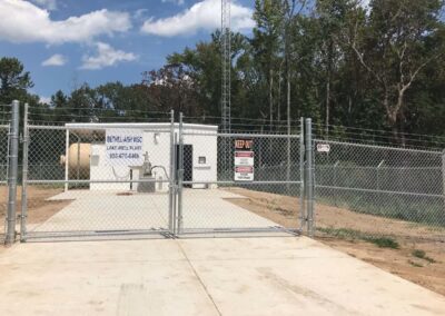 A fenced utility facility with warning signs, a white building, large tank, and security gate, surrounded by trees and grass under a blue sky with clouds.