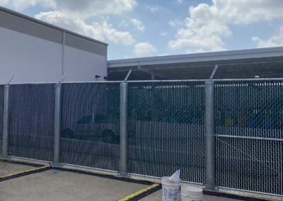 A chain-link fence with barbed wire on top stands next to a white building under a partly cloudy sky. Two white buckets sit on the ground near the fence.