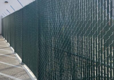A long chain-link fence with green privacy slats stands beside a concrete walkway. The fence casts diagonal shadows on the ground, and metal security spikes are attached along the top edge.