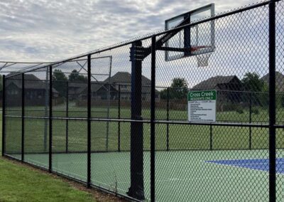 A fenced outdoor basketball court with a hoop and a sign on the fence that reads Cross Creek Residents Only. Houses and trees are visible in the background under a cloudy sky.