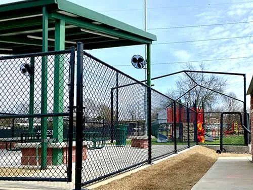 A chain-link fence surrounds a covered picnic area near a playground with colorful equipment on a sunny day; bare trees and blue sky are visible in the background.