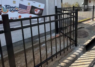 A black metal fence encloses a small, empty patch of dirt on a sidewalk near a street curb; behind the fence is a billboard displaying images of cards and the words Good Vibe.
