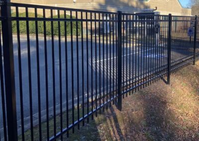 A black metal fence runs alongside a parking lot bordered by bushes, with a tan building and security cameras visible in the background on a sunny day.