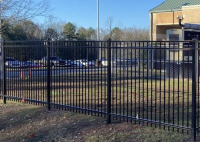 A black metal fence surrounds a grassy area near a building with a green roof. In the background, cars are parked along a street, and trees are visible beyond.
