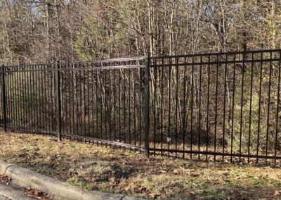 A metal fence with vertical bars stands on grass near a curb, with leafless trees and dense shrubs in the background on a sunny day.