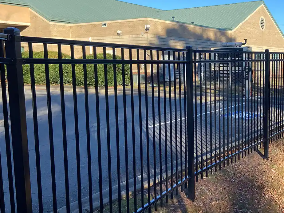A tall black metal fence with vertical bars encloses a paved parking lot in front of a large tan brick building with a green roof and trimmed bushes along the wall.