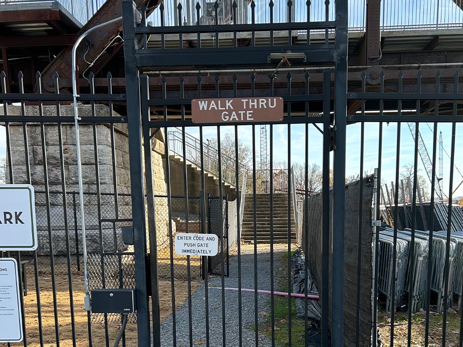 A black metal gate with a sign reading WALK THRU GATE, leading to a stairway behind the fence. A smaller sign instructs to ENTER CODE AND PUSH GATE IMMEDIATELY. Industrial structures are visible in the background.