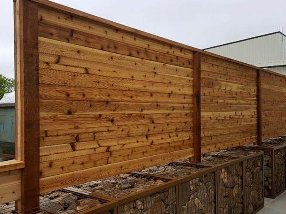 A tall wooden privacy fence with horizontal planks sits atop rectangular metal cages filled with stacked firewood, next to an industrial building.