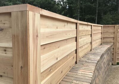 A close-up view of a modern wooden fence with horizontal planks, built alongside a curved brick wall. Green trees and foliage are visible in the background.